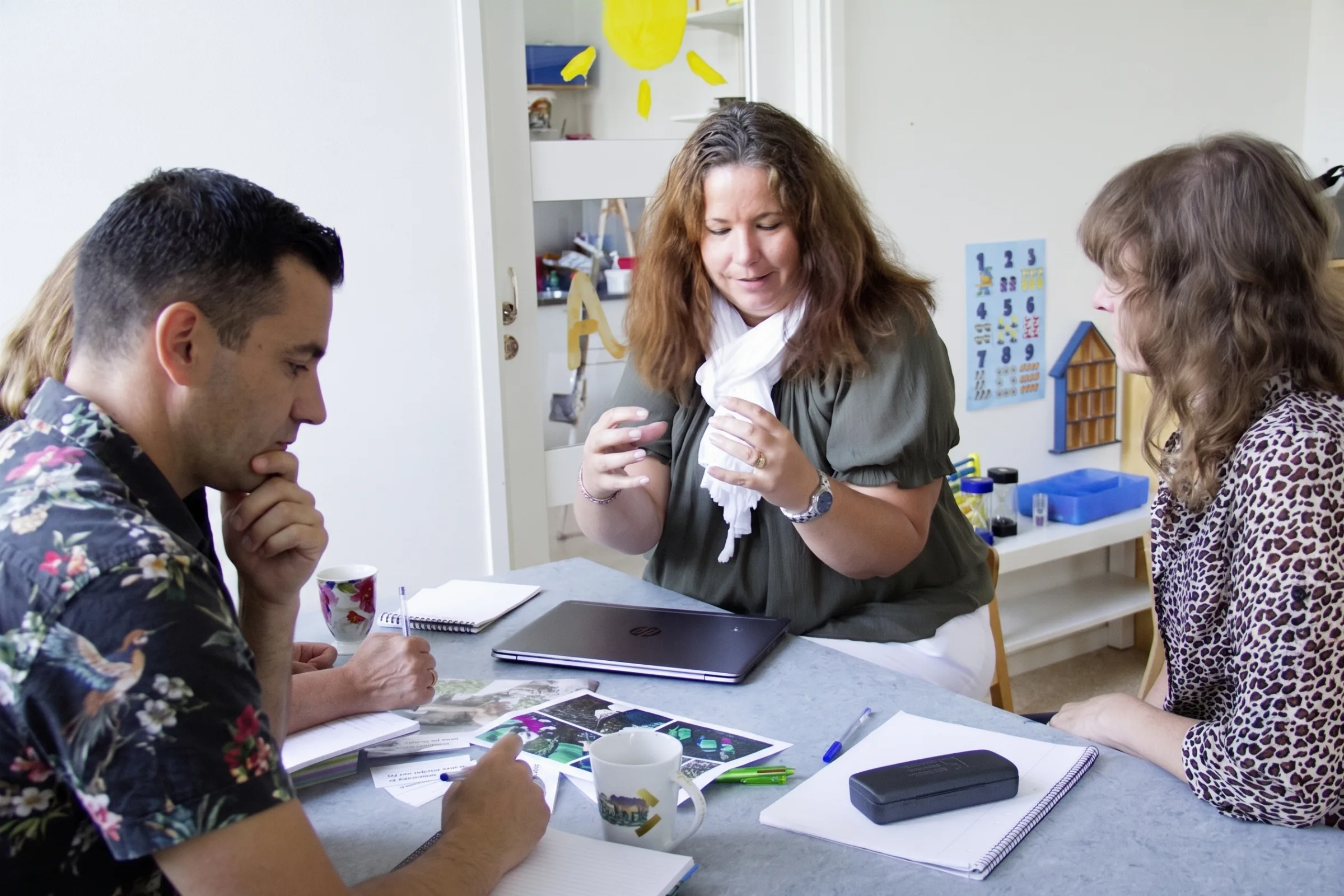 Three educators sit around a table in a preschool classroom, discussing materials and taking notes during a staff meeting.