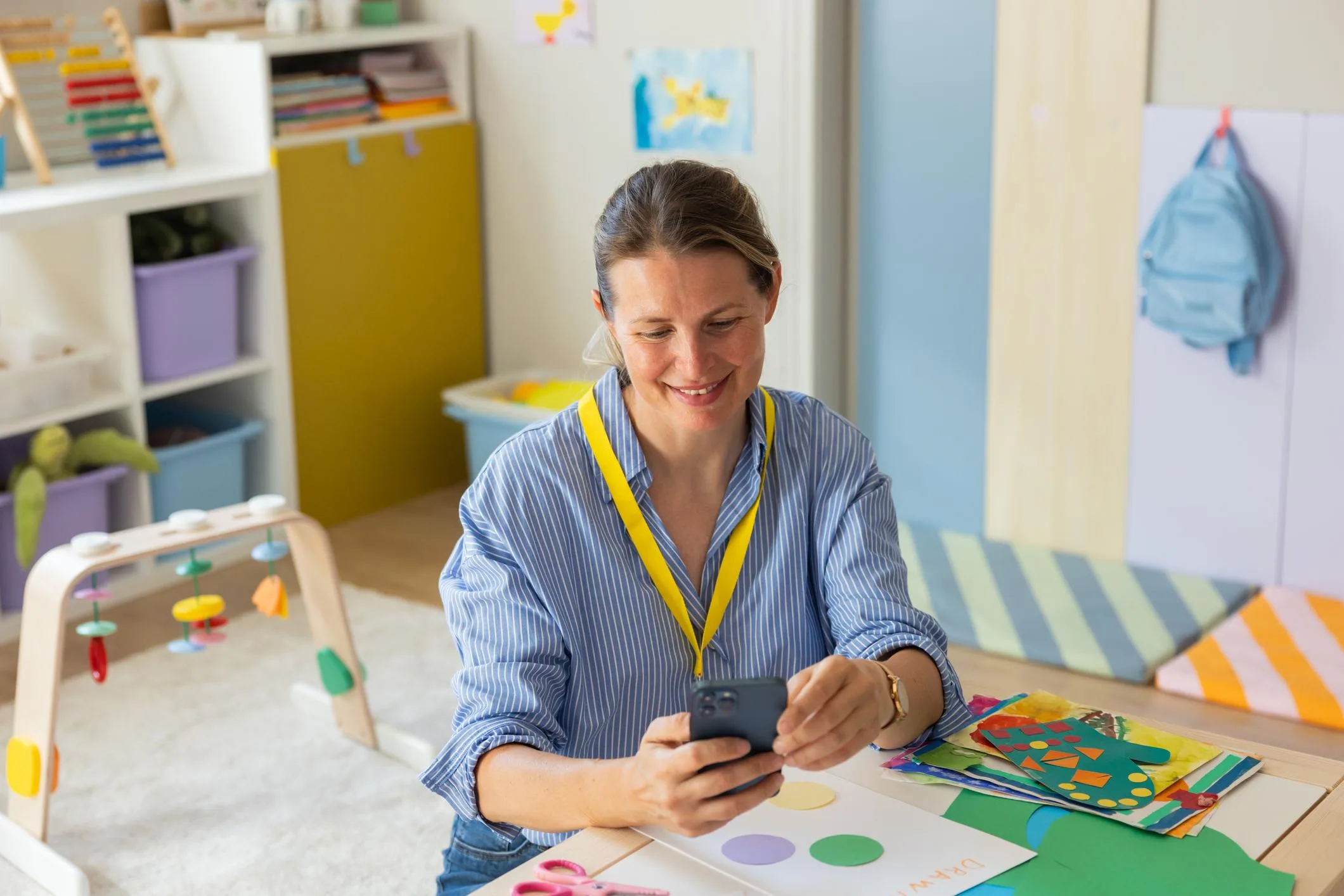 An educator seated in a preschool classroom looks at his smartphone, with children's artwork and classroom materials nearby.