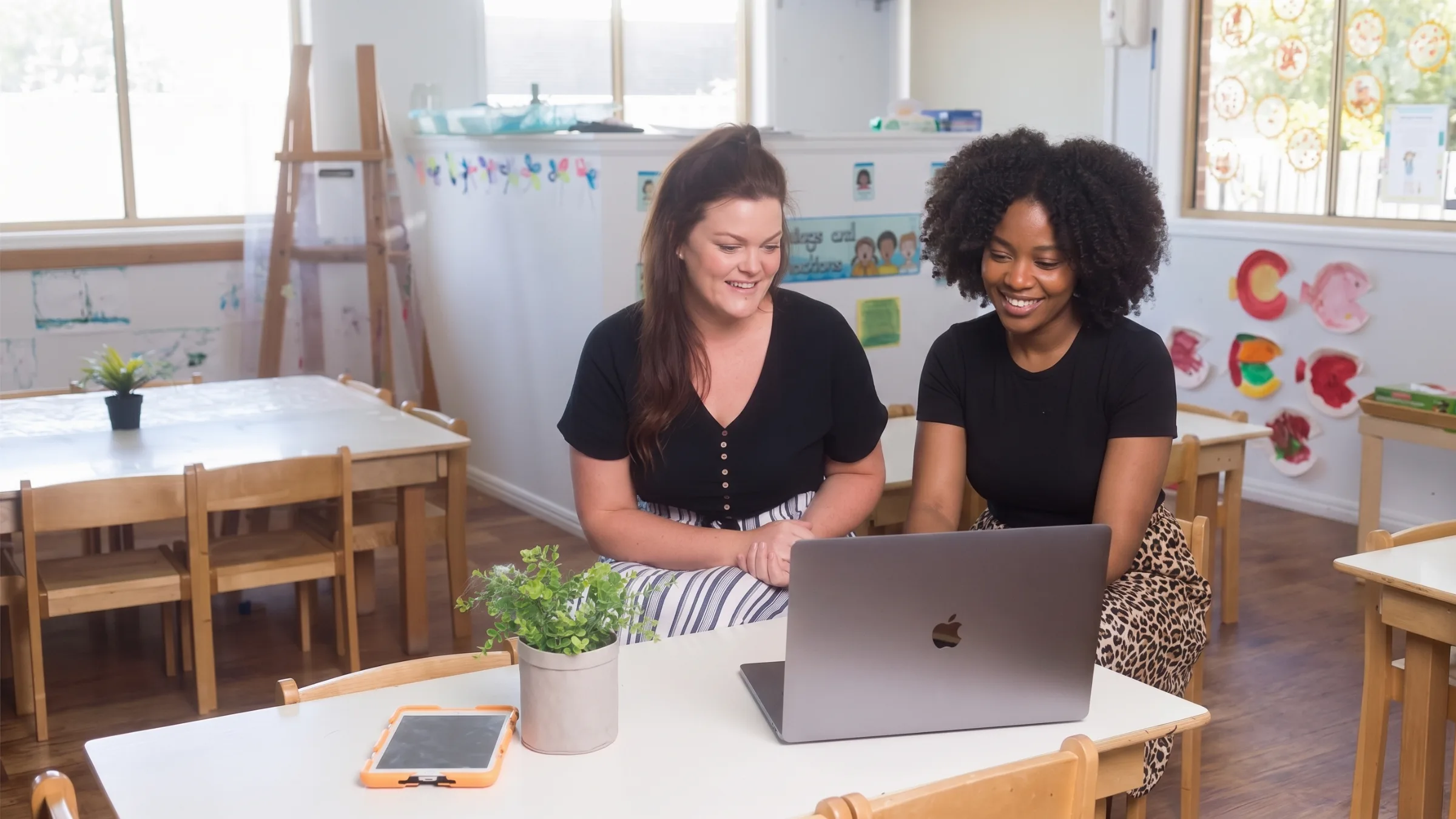 Two women sit together in a preschool classroom, smiling as they review a laptop on a child-sized table.